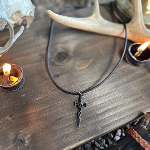 Może przedstawiać: Czarny naszyjnik z wisiorkiem w kształcie sztyletu. Urok ma skomplikowane szczegóły i wisi na cienkim, ciemnym sznurku. Obraz jest ustawiony na drewnianej powierzchni z zapalonymi świecami i dekoracyjnymi porożami.