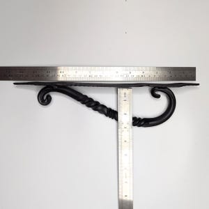 K&ouml;nnte beinhalten: Schwarze Metall-Regalhalterung mit gedrehtem Design und einer dekorativen Rolle an jedem Ende. Die Halterung ist etwa 20 cm lang und 7,5 cm breit.