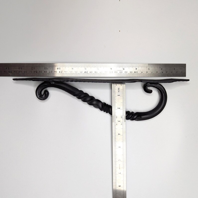 K&ouml;nnte beinhalten: Schwarze Metall-Regalhalterung mit gedrehtem Design und einer dekorativen Rolle an jedem Ende. Die Halterung ist etwa 20 cm lang und 7,5 cm breit.