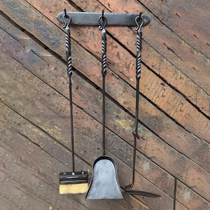 Può includere: Un set di attrezzi per camino in ferro battuto nero. Il set comprende un attizzatoio, una paletta, una spazzola e un supporto con volute decorative. Gli attrezzi hanno manici attorcigliati e sono appesi a una parete con assi di legno.