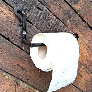 Może przedstawiać: Czarny, ręcznie kutego metalu uchwyt na papier toaletowy. Uchwyt jest zamontowany na rustykalnej drewnianej powierzchni i ma dekoracyjny kształt haka. Rolka białego papieru toaletowego jest na miejscu.