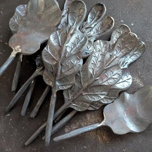 K&ouml;nnte beinhalten: Eine Sammlung handgefertigter Metall-Eichenblatt-Skulpturen. Jedes Blatt ist mit Adern versehen und an einem Stiel befestigt. Die Bl&auml;tter sind auf einer dunklen, strukturierten Oberfl&auml;che angeordnet, die die nat&uuml;rliche Farbe des Metalls zeigt.