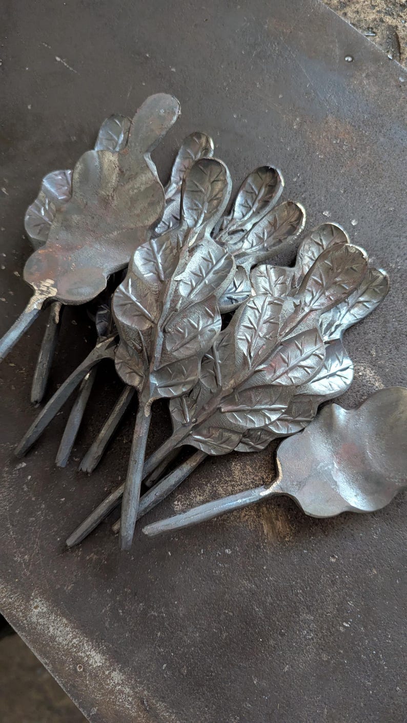 K&ouml;nnte beinhalten: Eine Sammlung handgefertigter Metall-Eichenblatt-Skulpturen. Jedes Blatt ist mit Adern versehen und an einem Stiel befestigt. Die Bl&auml;tter sind auf einer dunklen, strukturierten Oberfl&auml;che angeordnet, die die nat&uuml;rliche Farbe des Metalls zeigt.