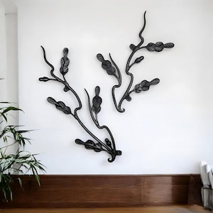 May include: Black metal wall art depicting stylized branches with leaves. The art is mounted on a white wall, with a dark wooden surface below. The branches have a flowing, organic design, adding a touch of nature-inspired decor.