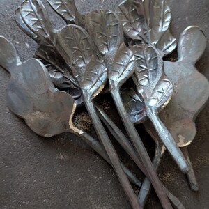 K&ouml;nnte beinhalten: Eine Sammlung handgefertigter Metallkunstwerke mit blattf&ouml;rmigen Designs. Die St&uuml;cke haben eine dunkle, metallische Oberfl&auml;che und sind auf einer strukturierten Oberfl&auml;che angeordnet. Die Bl&auml;tter haben detaillierte Adern.