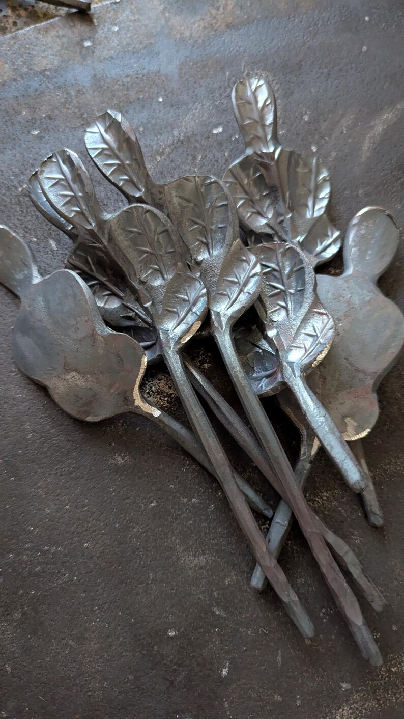 K&ouml;nnte beinhalten: Eine Sammlung handgefertigter Metallkunstwerke mit blattf&ouml;rmigen Designs. Die St&uuml;cke haben eine dunkle, metallische Oberfl&auml;che und sind auf einer strukturierten Oberfl&auml;che angeordnet. Die Bl&auml;tter haben detaillierte Adern.