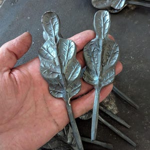 K&ouml;nnte beinhalten: Nahaufnahme mehrerer handgefertigter Metallblatt-Skulpturen. Zwei werden in einer Hand gehalten und zeigen detaillierte Adern und eine strukturierte Oberfl&auml;che. Die Bl&auml;tter sind dunkelgrau, mit langen Stielen. Der Hintergrund zeigt weitere Blatt-Skulpturen.