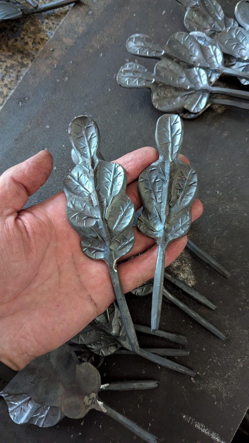 K&ouml;nnte beinhalten: Nahaufnahme mehrerer handgefertigter Metallblatt-Skulpturen. Zwei werden in einer Hand gehalten und zeigen detaillierte Adern und eine strukturierte Oberfl&auml;che. Die Bl&auml;tter sind dunkelgrau, mit langen Stielen. Der Hintergrund zeigt weitere Blatt-Skulpturen.