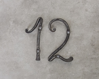 May include: A metal house number plaque with the number "12" in a decorative, stylized font. The number is attached to a light gray wall.