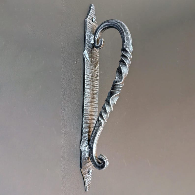 K&ouml;nnte beinhalten: Ein schwarzer T&uuml;rgriff aus Metall mit einem gedrehten Design. Der Griff ist an einer rechteckigen Metallplatte mit einem dekorativen Design befestigt.
