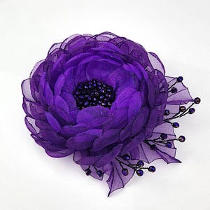 Può includere: Un broche a forma di fiore in tessuto viola con un centro di perline nere. Il fiore ha un design a strati e arricciato.