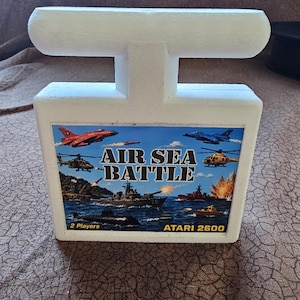 May include: A white Atari 2600 game cartridge holder featuring a rectangular label with the title Air Sea Battle. The label displays a colorful scene of military aircraft, ships, and a submarine in combat, with the text 2 Players and Atari 2600.