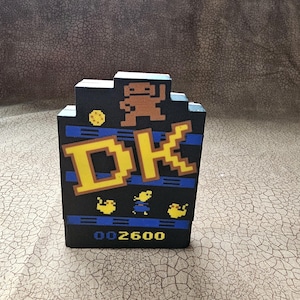 May include: A black rectangular video game cartridge with a pixelated graphic of a brown ape, yellow letters reading DK, and small yellow and blue characters. The bottom of the cartridge features the blue and yellow text 002600 on a black background.