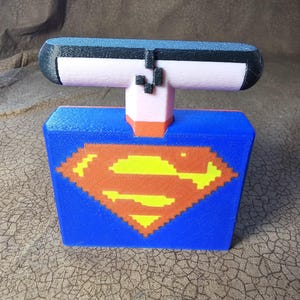 May include: A 3D printed figure featuring a blue rectangular base with a pixelated yellow and orange Superman-style emblem on the front. The figure has a light pink neck section and a horizontal black top piece with a small black pixelated detail.