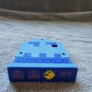 May include: A blue 3D-printed display stand featuring a light blue ghost-shaped top section. The front base displays the text RetroCartArt in blue, with two purple ghost icons and a yellow circular character icon printed on the lower front edge.