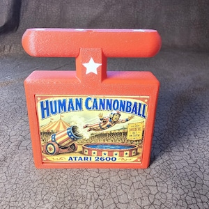 May include: A bright orange plastic Atari 2600 game cartridge holder featuring a rectangular base and a T-shaped top. The front displays a colorful graphic of a human cannonball act with the text Human Cannonball and Atari 2600 printed clearly.
