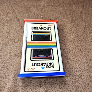 May include: An Atari 2600 Super Breakout video game cartridge with a white label featuring black text, a rainbow stripe, and two game screen illustrations. The cartridge has a bright orange top edge and a blue bottom edge with ribbed plastic.