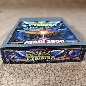 May include: A black Atari 2600 video game cartridge for the game Phoenix. The label features a colorful space scene with a spaceship, planets, and the text Phoenix and Atari 2600. The end label also displays the title Phoenix with wings.