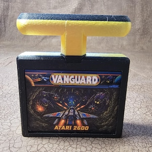 May include: A black Atari 2600 video game cartridge for Vanguard with a yellow T-shaped handle attached to the top. The label features a colorful space combat illustration with the text Vanguard and Atari 2600 printed in white and yellow.