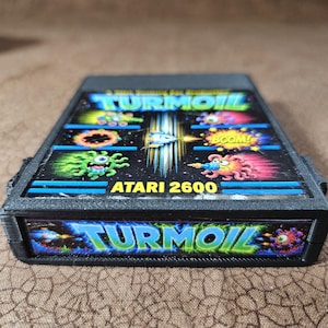 May include: A black Atari 2600 video game cartridge for the game Turmoil. The label features colorful graphics of cartoon creatures, a spaceship, and the text Turmoil and Atari 2600. The end label also displays the game title Turmoil in blue text.