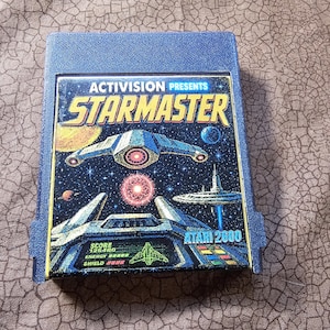 May include: A blue Atari 2600 video game cartridge for the game Starmaster by Activision. The label features a space scene with a starship, planets, and a cockpit view displaying score, energy, and shield levels in a pixelated graphic style.
