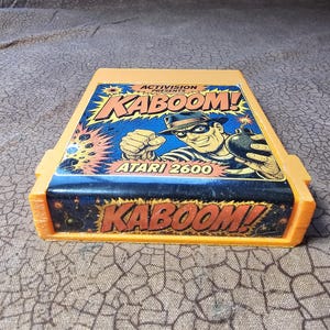 May include: An orange Atari 2600 video game cartridge for the game Kaboom! by Activision. The label features a comic-style illustration of a masked character holding a bomb, with the text Activision Presents Kaboom! and Atari 2600 visible.