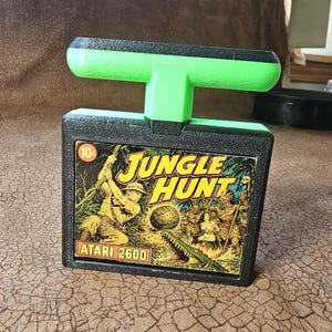 May include: A black and neon green Atari 2600 game cartridge handle featuring a Jungle Hunt label. The rectangular label displays a jungle scene with a person swinging on a vine, a crocodile, and the text Jungle Hunt and Atari 2600.