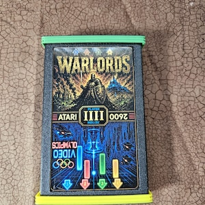 May include: A rectangular Atari 2600 video game cartridge for Warlords and Video Olympics. The black plastic cartridge features a label with a knight illustration, the text Warlords, Atari, and Video Olympics, and bright green plastic end caps.