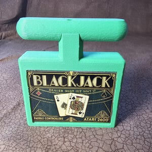 May include: A bright green Atari 2600 Blackjack paddle controller housing with a rectangular base and a top handle. The front features a black label with the text BLACKJACK, DEALER MUST HIT SOFT 17, PADDLE CONTROLLERS, and ATARI 2600.