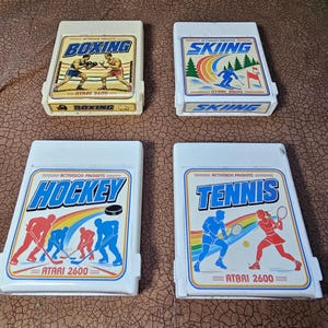 May include: Four white Atari 2600 video game cartridges by Activision. The labels feature colorful illustrations and the titles Boxing, Skiing, Hockey, and Tennis. Each cartridge displays the Activision Presents logo and the Atari 2600 console name.