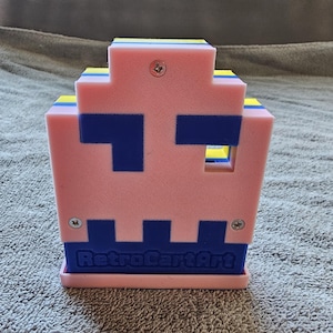 May include: A 3D-printed pixelated ghost figure in light pink and dark blue with yellow accents. The front features a cutout eye and the embossed text RetroCartArt at the base. Three silver metal screws are visible on the front surface of the object.
