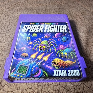 May include: A purple Atari 2600 video game cartridge for the game Spider Fighter. The label features a blue tank firing lasers at various colorful, multi-eyed spider creatures against a dark space background with a planet and stars.