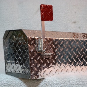 May include: A silver diamond-plate mailbox with a red signal flag. The mailbox has a rectangular shape with a sloped top and is made of a durable, textured metal. The red flag is attached to the side.