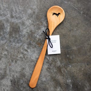 Puede incluir: Una cuchara de madera con una silueta de dachshund tallada en el cuenco. La cuchara est&aacute; atada con una cuerda negra y una etiqueta blanca que dice "Kuchen Helfer Seesucht".