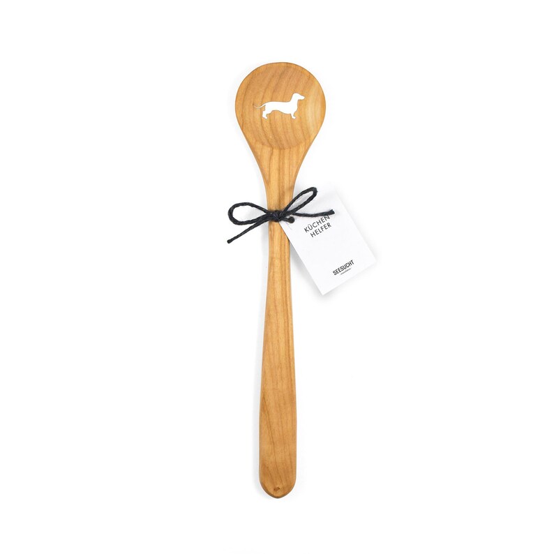 Puede incluir: Una cuchara de madera con un dise&ntilde;o de dachshund recortado. La cuchara est&aacute; hecha de madera natural y tiene una cuerda negra atada alrededor del mango con una etiqueta blanca que dice "Kuchen Helfer" y "Seesucht".