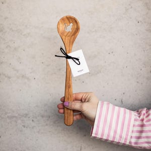 May include: Wooden spoon with a cat-shaped cutout in the bowl. The spoon is tied with black twine and has a white tag attached. The spoon is held by a person wearing a pink and white striped shirt.