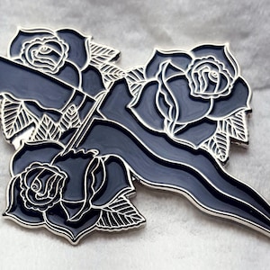 May include: Black enamel pin featuring a stylized dagger with three black roses. The roses have white outlines and are arranged around the dagger.