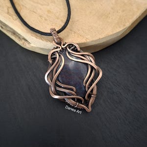 May include: A pendant featuring a dark, mottled purple and black stone encased in intricate, swirling copper wire wrapping. The pendant hangs from a simple black cord and includes the text Danea Art printed in small white letters below it.
