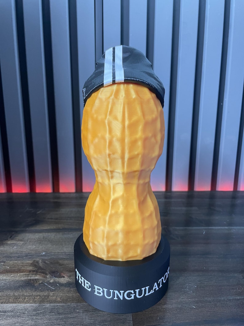 May include: A whimsical, 3D-printed peanut sculpture wearing a black cap with white stripes. The peanut is tan with a textured surface, mounted on a black base with the text "THE BUNGULATOR".