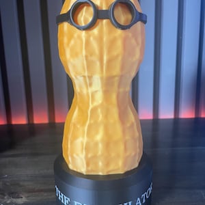 May include: A 3D-printed sculpture of a peanut wearing black glasses, mounted on a black base. The base has the text "THE BUNGULATOR" in white. The peanut is a light orange color.