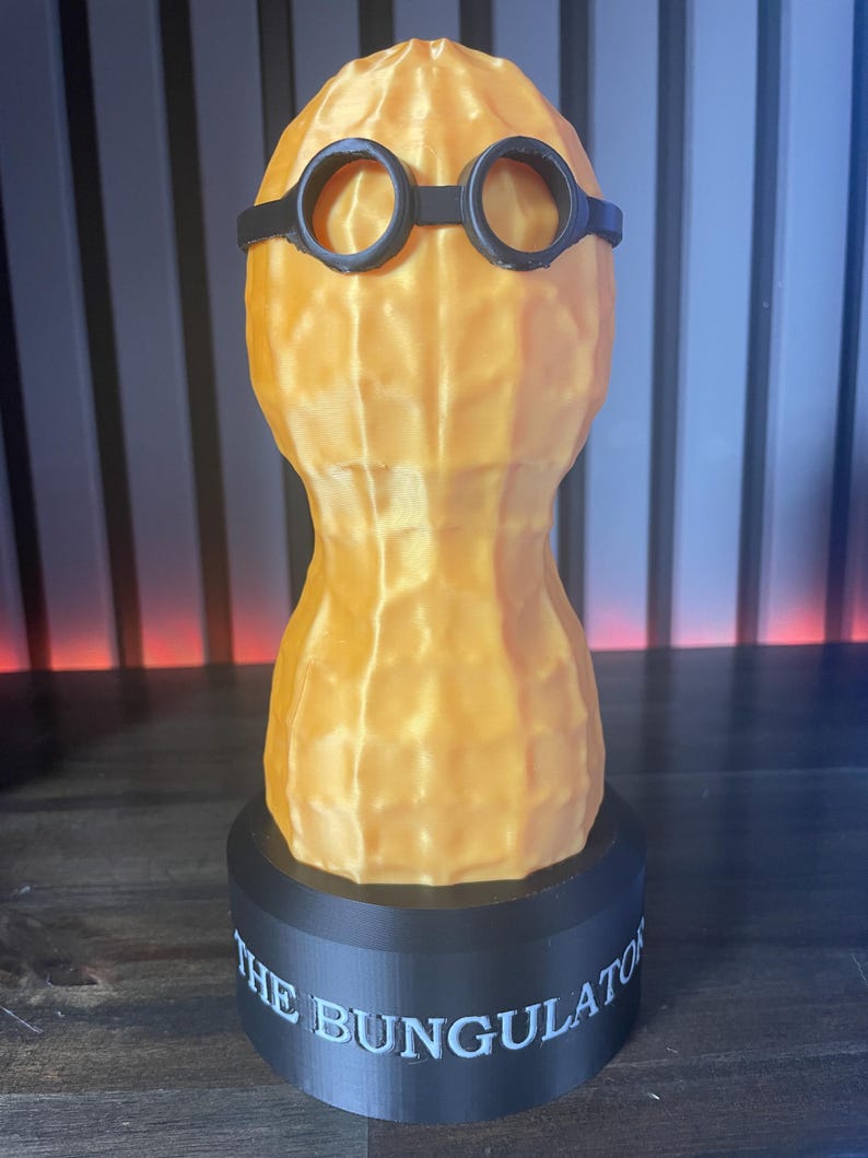 May include: A 3D-printed sculpture of a peanut wearing black glasses, mounted on a black base. The base has the text "THE BUNGULATOR" in white. The peanut is a light orange color.