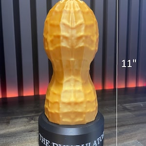 May include: A golden-colored, peanut-shaped sculpture, 11 inches tall, sits atop a black base. The base is inscribed with the text "THE BUNGULATOR". The sculpture has a textured surface, resembling a peanut shell.