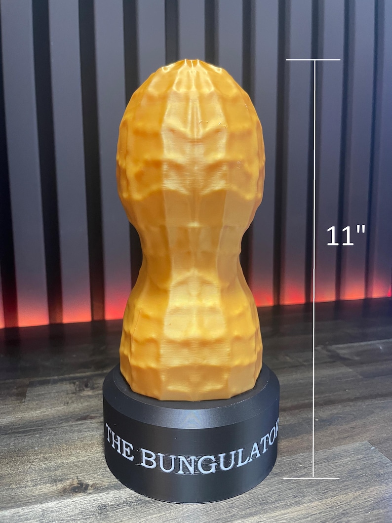 May include: A golden-colored, peanut-shaped sculpture, 11 inches tall, sits atop a black base. The base is inscribed with the text "THE BUNGULATOR". The sculpture has a textured surface, resembling a peanut shell.