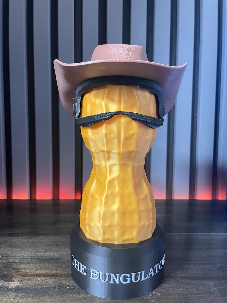 May include: A 3D-printed sculpture of a peanut wearing a pink cowboy hat and black sunglasses. The peanut is orange and textured, mounted on a black base with the text "THE BUNGULATOR".