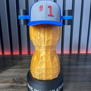 May include: A novelty sculpture of a peanut with a baseball cap that has the number 1 on it. The peanut is tan and sits on a black base with the words "THE BUNGULATOR" printed on it. Two black cylinders are attached to the sides of the cap.