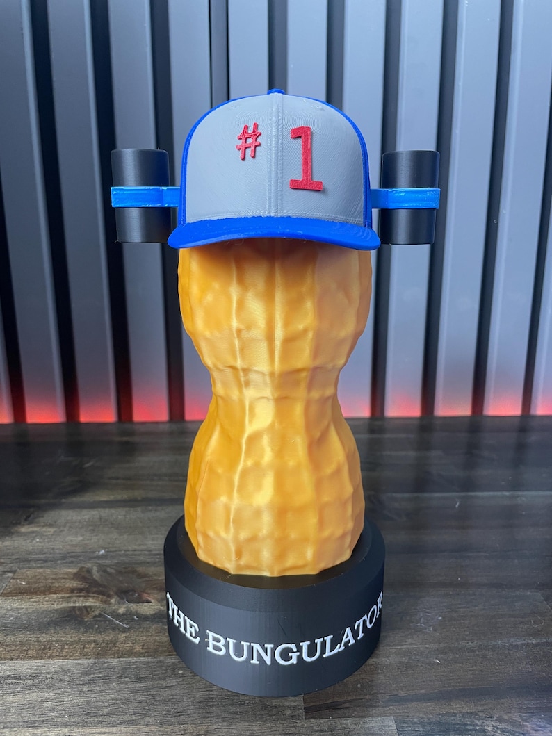 May include: A novelty sculpture of a peanut with a baseball cap that has the number 1 on it. The peanut is tan and sits on a black base with the words "THE BUNGULATOR" printed on it. Two black cylinders are attached to the sides of the cap.