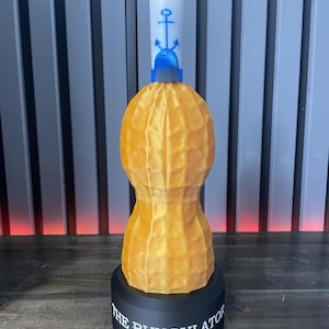 May include: A 3D-printed novelty item shaped like a peanut, with a white top featuring a blue anchor design. The peanut is a textured, golden-yellow color, and it sits on a black base with the text "THE BUNGULATOR".