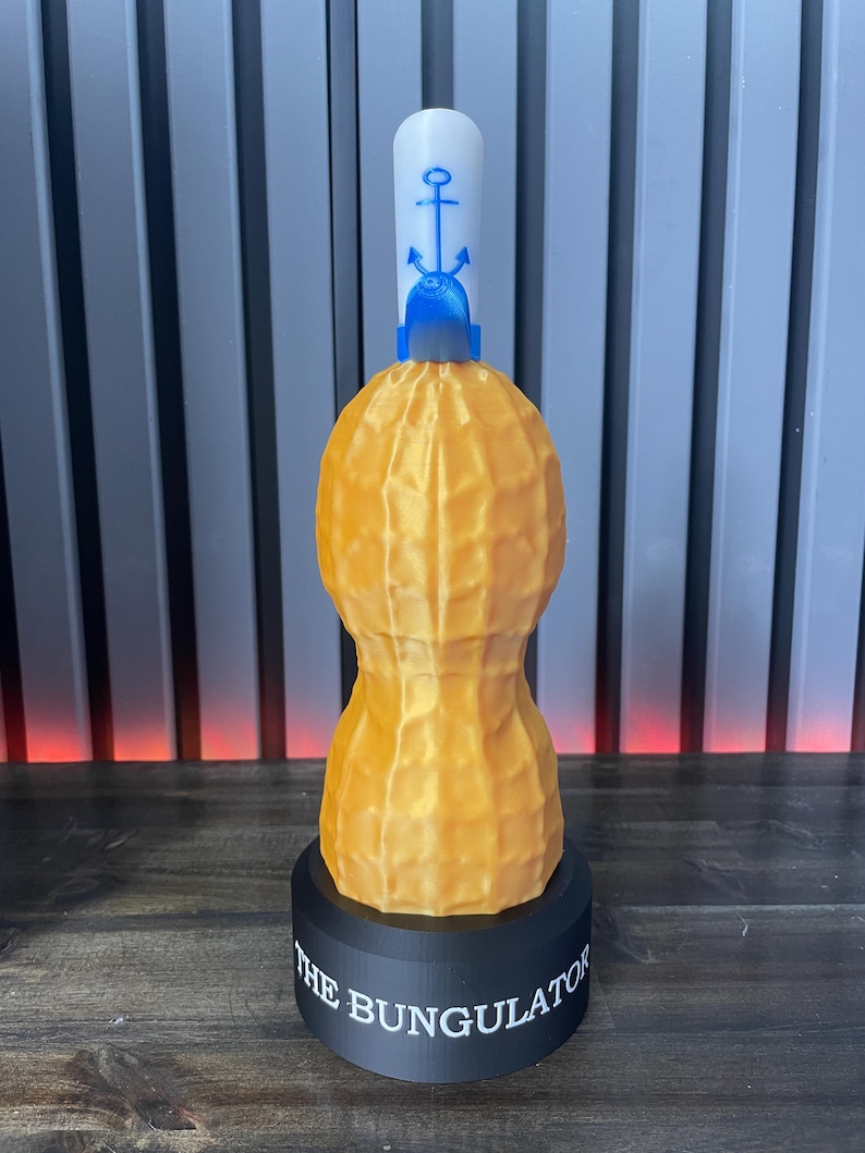 May include: A 3D-printed novelty item shaped like a peanut, with a white top featuring a blue anchor design. The peanut is a textured, golden-yellow color, and it sits on a black base with the text "THE BUNGULATOR".