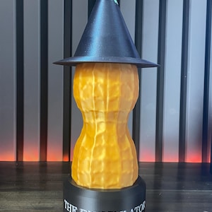 May include: A whimsical, 3D-printed sculpture of a peanut wearing a witch's hat. The peanut is textured and golden yellow, with a black hat and a green tip. The base is black with the text "THE BUNGULATOR".