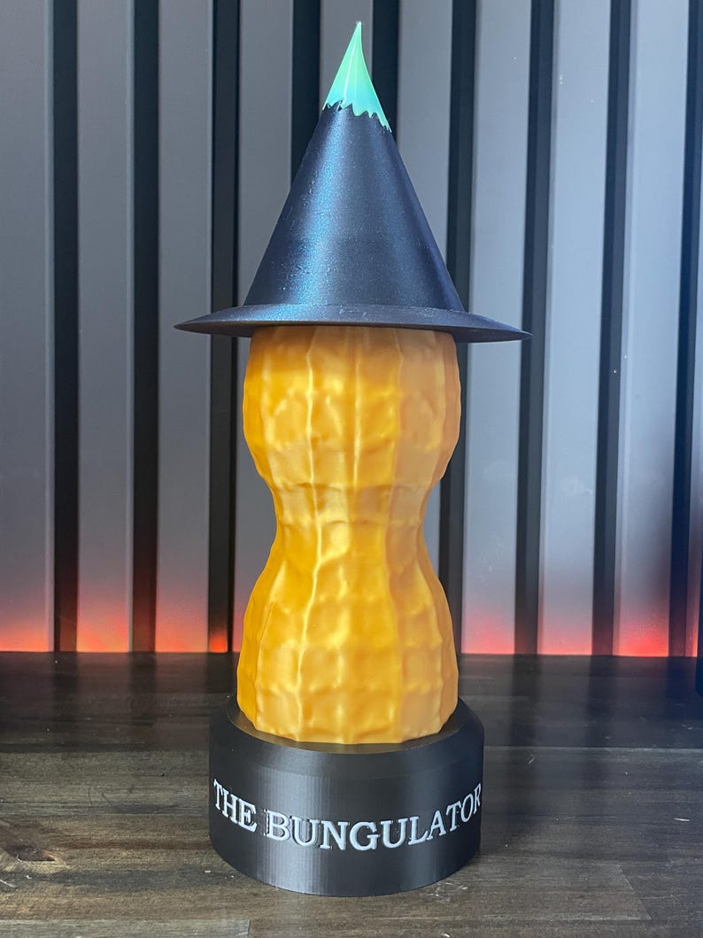 May include: A whimsical, 3D-printed sculpture of a peanut wearing a witch's hat. The peanut is textured and golden yellow, with a black hat and a green tip. The base is black with the text "THE BUNGULATOR".